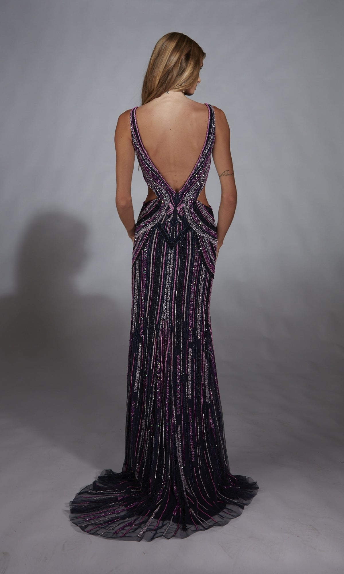 A woman faces away, wearing the Alyce 61892 long prom dress featuring a sleeveless, floor-length open-back design with intricate shimmering purple and silver beadwork.